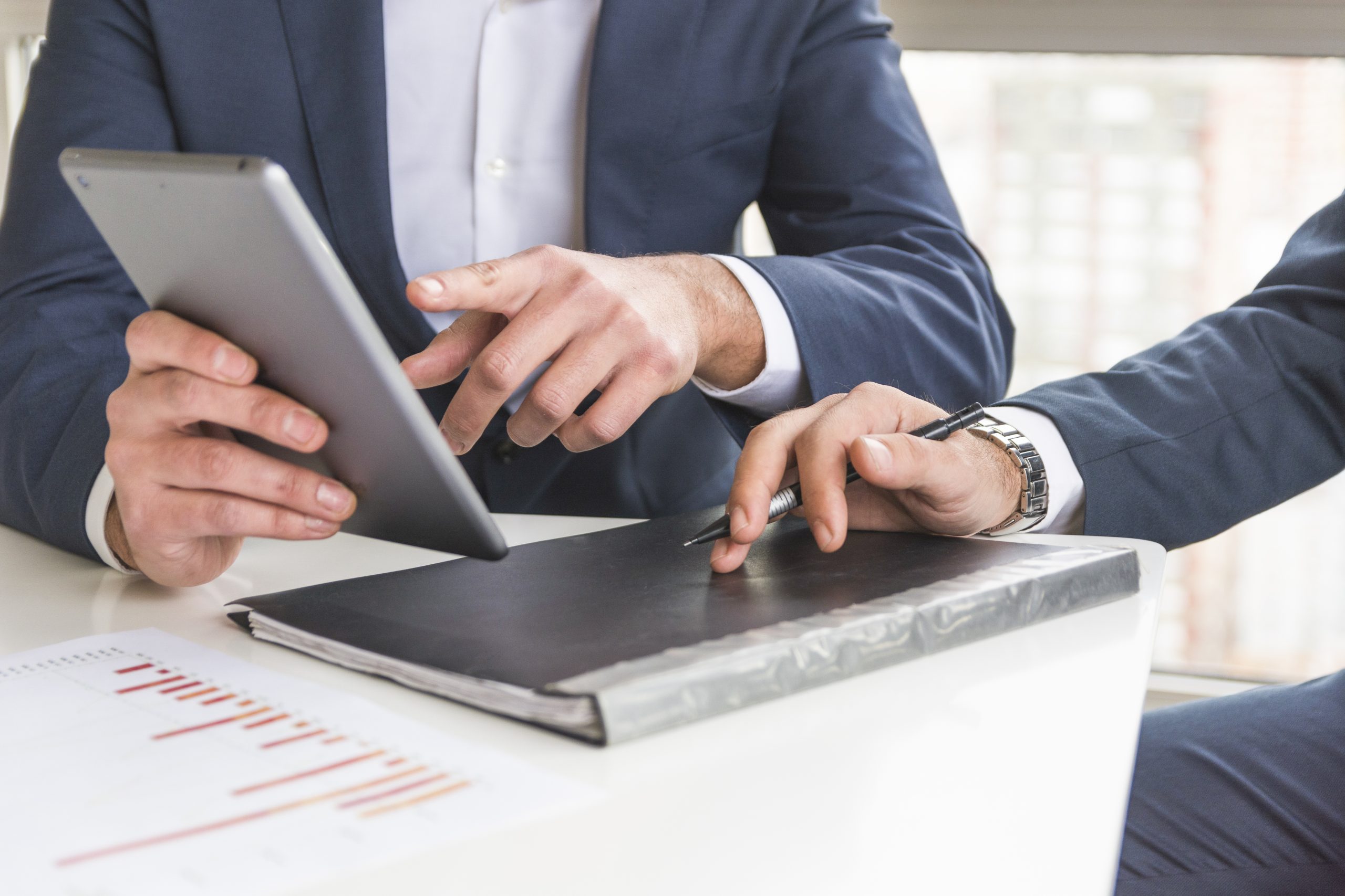 Two businessmen in formal attire discussing business documents and financial data using a digital tablet and printed reports